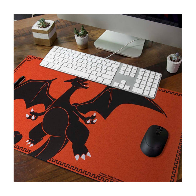 Playmat premium de Charizard con diseño de llamas, borde bordado y base antideslizante con patrón de Poké Ball. Imagen con una computadora. 